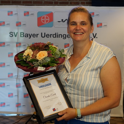 Frau mit Blumenstrauß und Urkunde in der Hand, lächelnd vor einem Banner des SV Bayer Uerdingen e.V.