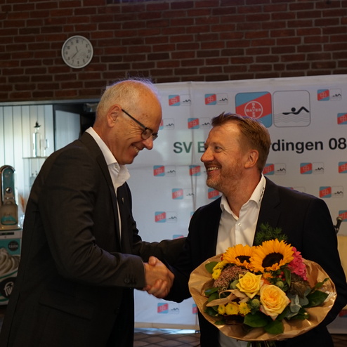 Zwei Männer schütteln sich die Hände, einer hält einen Strauß mit bunten Blumen in der anderen Hand.