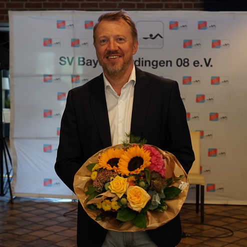 Mann in Anzug hält einen bunten Blumenstrauß vor einem Veranstaltungsbanner des SV Bayreuth 08 e.V. im Hintergrund.
