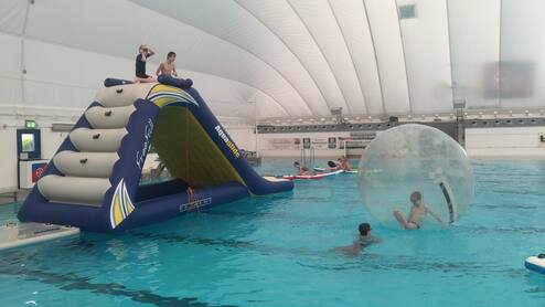 Kinder spielen auf einem großen aufblasbaren Spielgerät im Wasser, andere schwimmen in durchsichtigen Wasserbällen.