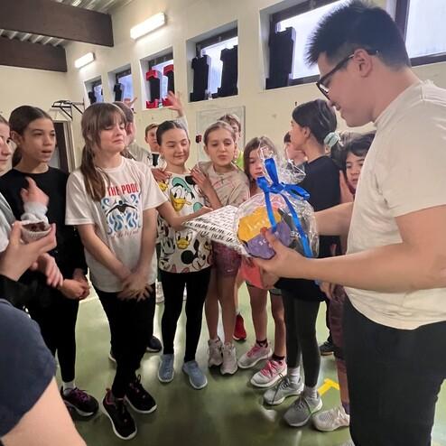 Gruppe von Kindern schaut gespannt auf ein Geschenk, das von einem Erwachsenen in einem Sportraum gehalten wird.