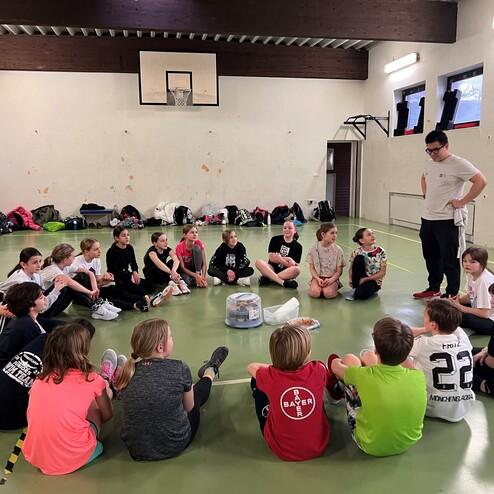 Gruppensitzung mit Kindern im Sportraum, umgeben von Rucksäcken; ein Trainer steht und spricht.