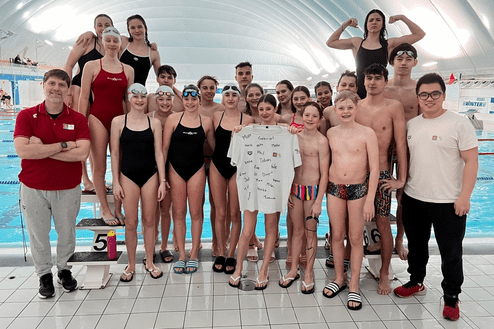 Gruppenfoto von Schwimmern und Trainern im Hallenbad, mit einem T-Shirt in der Mitte, das persönliche Botschaften trägt.