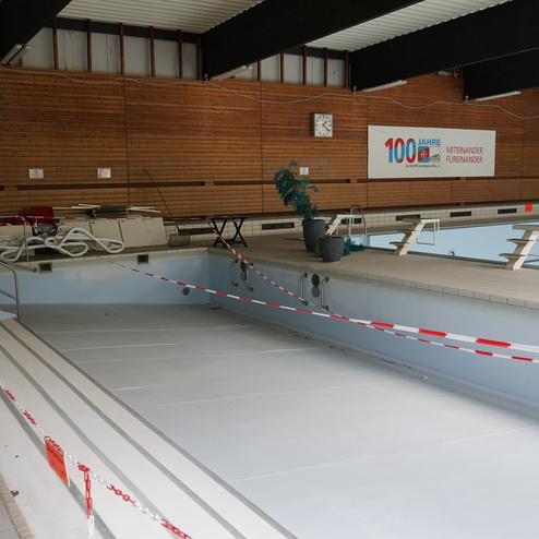 Leerer Schwimmbereich mit Absperrband, Liegen und einem Banner zum Jubil&auml;um an der Wand.