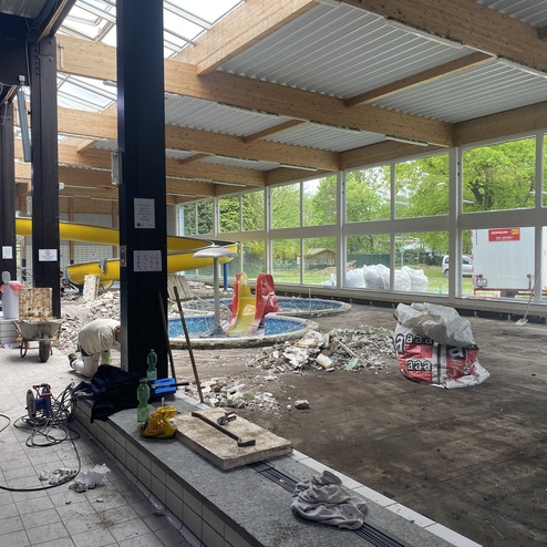 Baustelle in einem Schwimmbad mit einem Kinderrutschenbereich, einem leeren Pool und Baustellenmaterialien im Innenbereich.