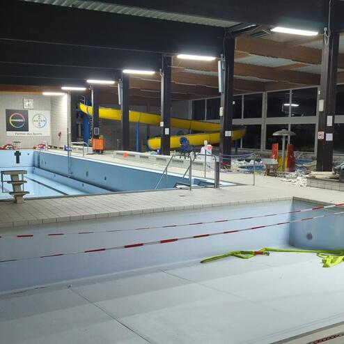 Leeres Schwimmbad mit zwei Sprungtürmen, einer Rutsche und Sicherheitsausrüstung, umgeben von Baustellenmaterial.