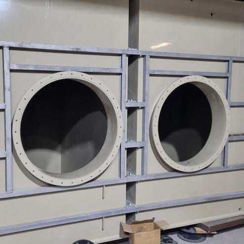 Graue industrielle Struktur mit zwei runden Öffnungen, Metallrahmen und Rohren im Innenbereich einer Werkhalle.