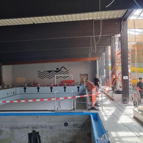 Blick auf ein Schwimmbad im Umbau mit leeren Becken und Bauarbeitern, einer gelben Rutsche und Sicherheitsbarrieren.