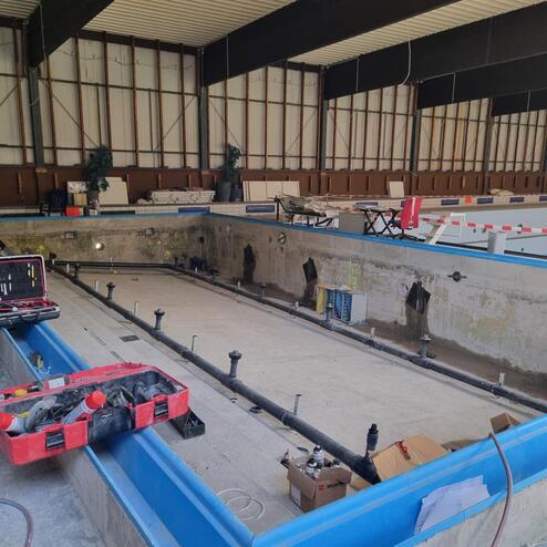 Blick auf einen leeren Betonpool mit Rohren, Werkzeugen und Baustellenmaterial in einer unvollendeten Schwimmhalle.