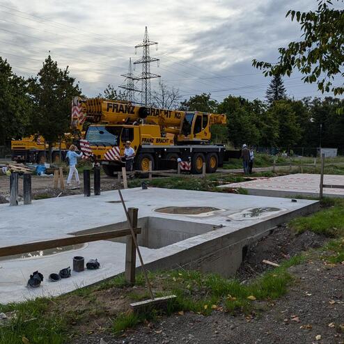 Baustelle mit Kränen, Arbeiter und Betonfundamenten in einer grünen Umgebung unter bewölktem Himmel.