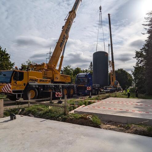 Bauarbeiter positionieren einen großen Zylinder mit Kränen auf einem Baustellenareal zwischen Bäumen.