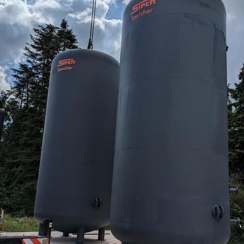 Zwei große, graue Speichertanks stehen auf einer Betonfläche, umgeben von Bäumen und teilweise bewölktem Himmel.