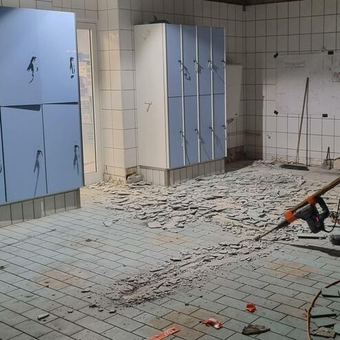 Arbeiter entfernen Fliesen in einem Umkleideraum mit blauen Spinden und unordentlicher Baustelle.