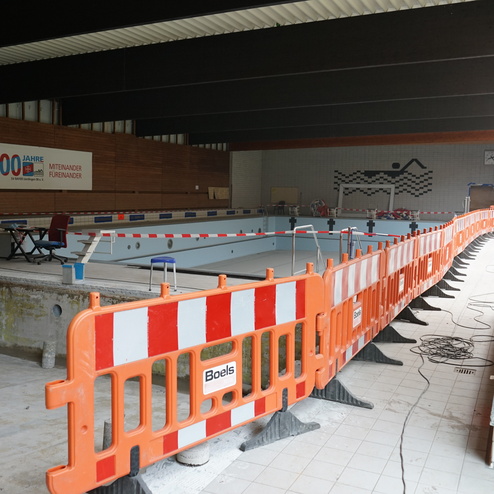 Sanierungsarbeiten in einem Schwimmbad, mit Absperrungen, Baustelle und einem Teil des leeren Schwimmbeckens.