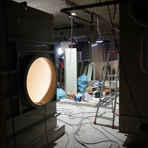 Baustelle mit gro&szlig;en Ger&auml;ten im Hintergrund, Leiter, Werkzeug und Kabel auf dem Boden, schwaches Licht aus mehreren Quellen.