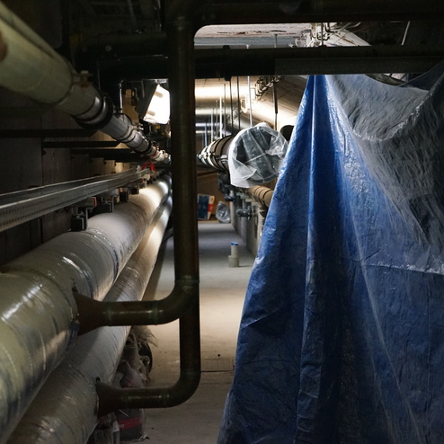 Enge Installationspassage mit Rohren, einem blauen Sichtschutz und schwachem Licht im Hintergrund.