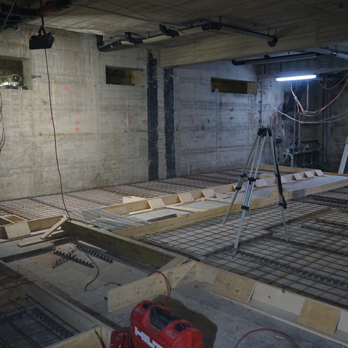Blick in einen Rohbau mit Metallbewehrung, Holzschalungen und Baustellenbeleuchtung. Kabel h&auml;ngen von der Decke.