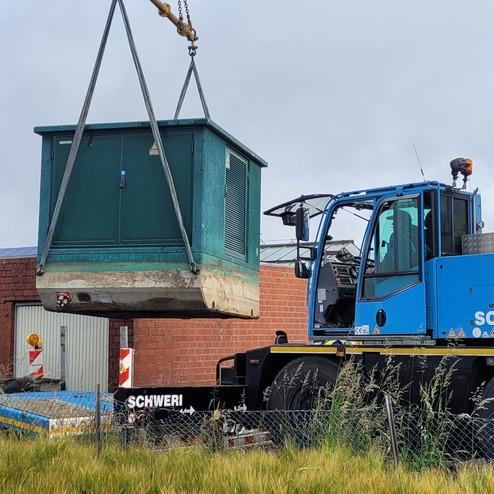 Kran hebt einen grünen containerartigen Bau auf einer Baustelle, im Hintergrund ist ein rotes Mauerwerk sichtbar.