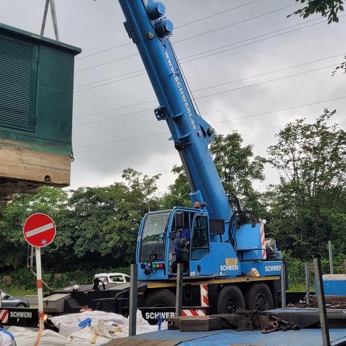 Lkw mit Kran hebt einen elektrischen Transformator über einer Baustelle, um ihn auf einen Tieflader zu platzieren.