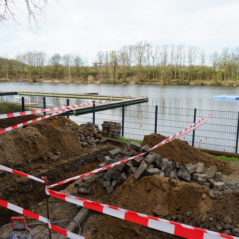 Baustelle an einem Gew&auml;sser mit Umgebung, Baustellenabsperrung, Erdarbeiten und haufenweise Steinen im Vordergrund.