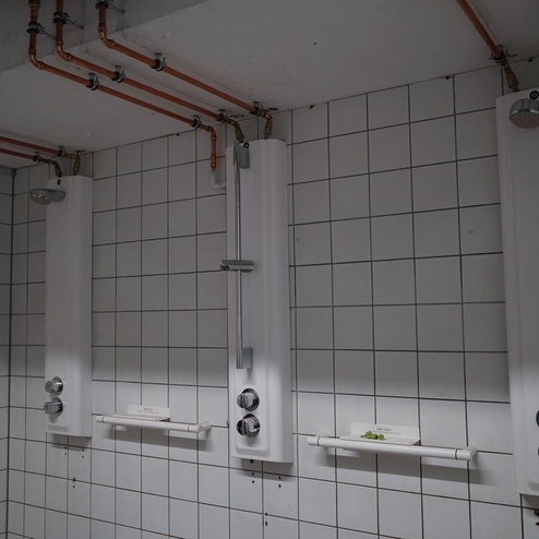 Drei Duschpaneele mit Armaturen und Haltegriffen in einem gefliesten Raum mit sichtbaren Wasserleitungen.