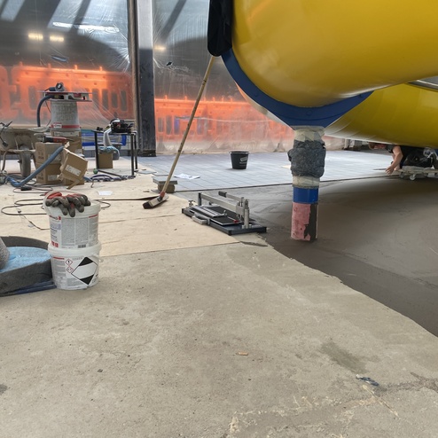 Blick auf Baustelle mit gelben Rohr, glattem Boden und Werkzeugen, w&auml;hrend eine Person den Boden bearbeitet.