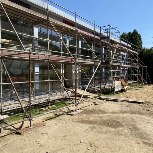 Blick auf ein Geb&auml;ude mit Ger&uuml;st und Fenstern, umgeben von unbefestigtem Boden und Gr&uuml;nfl&auml;chen.