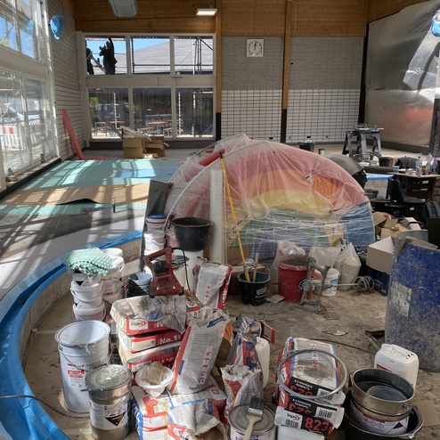 Baustelle mit verschiedenen Farbcontainern, einem bunten, eingeh&uuml;llten Objekt und handwerklichen Werkzeugen im Hintergrund.