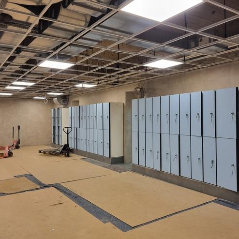 Innenansicht eines Umkleideraums mit blauen Spinden, Holz- und Linoleumfußboden sowie handgeführten Transportgeräten.