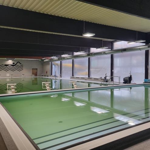 Blick auf ein Hallenbad mit grünem Wasser, Sprungbrettern und großen Fenstern, durch die Tageslicht fällt.