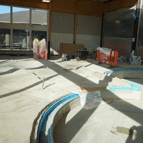 Bauinterieur mit unvollendetem Boden, geschwungenen Betonkanten und Baustellenmaterialien im Sonnenlicht.