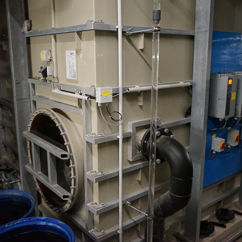 Technische Anlage mit gro&szlig;em Beh&auml;lter, Rohren und Schaltk&auml;sten in einer Industrieumgebung, umgeben von blauen F&auml;ssern.