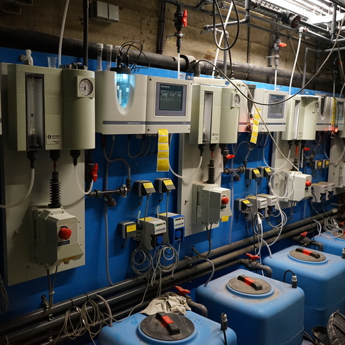 Technischer Raum mit mehreren Messger&auml;ten, Steuerungen und blauen Tanks an einer Wand. Kabel und Schl&auml;uche verlaufen sichtbar.