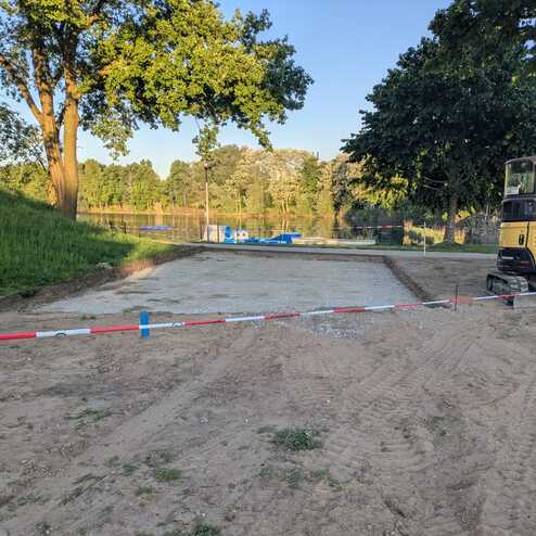 Baustelle am Ufer mit abgegrenztem Schotterbereich, umgeben von Bäumen und Blick auf einen ruhigen See.