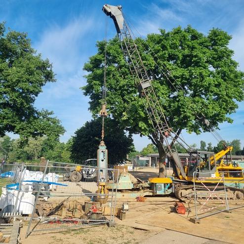 Bauplatz mit einem Kran, umgeben von Baustellenmaterial und Bäumen unter blauem Himmel.