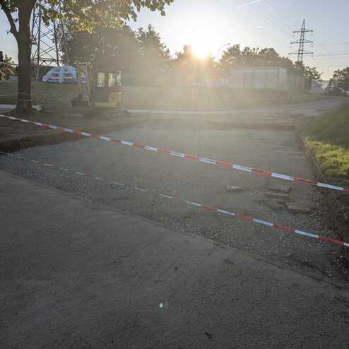 Bauarbeiten an einer Straße mit Absperrband, einem Bagger und Sonnenuntergang im Hintergrund.