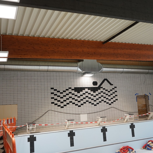 Innenansicht eines Schwimmbades mit Fliesenwand, Sicherheitsabsperrung und grafischem Motiv eines Schwimmers.