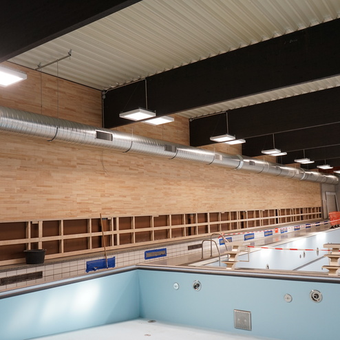 Innenansicht eines leerstehenden Schwimmbeckens mit Holz- und Metallakzenten sowie sichtbaren Deckenleuchten und L&uuml;ftungsrohren.