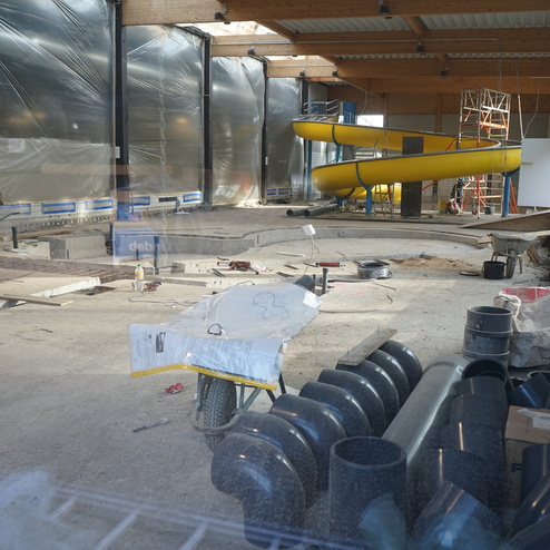 Bauarbeiten in einem Innenbereich mit gelben Rutsche, Rohren und Baustellenmaterialien auf dem Boden.