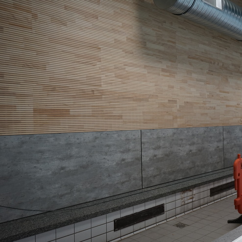 Holzwand mit horizontalen Leisten, darunter graue W&auml;nde und eine lange, grau gemusterte Ablagefl&auml;che. Sicherheitsabsperrung in Orange.