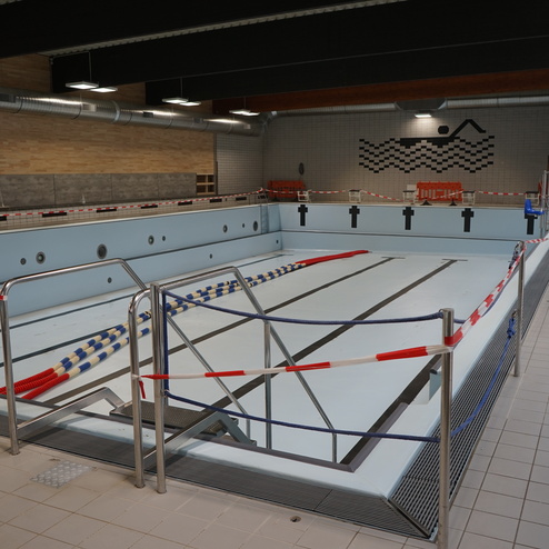 Leeres Schwimmbecken mit Absperrband und Liegen in einer Innenansicht eines Schwimmbads.