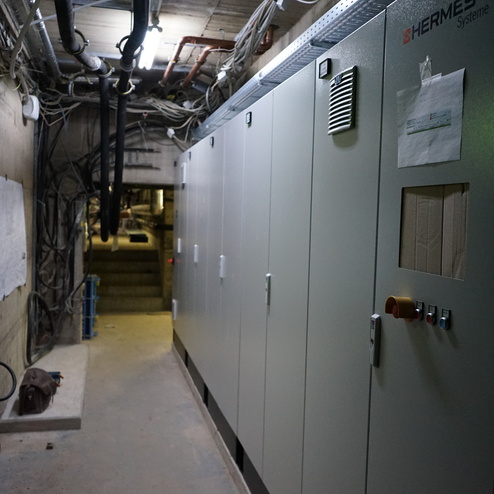 Dunkler Gang mit mehreren Schaltschr&auml;nken und Kabeln an der Wand, sowie einer Leiter und einer Feuerl&ouml;scher.