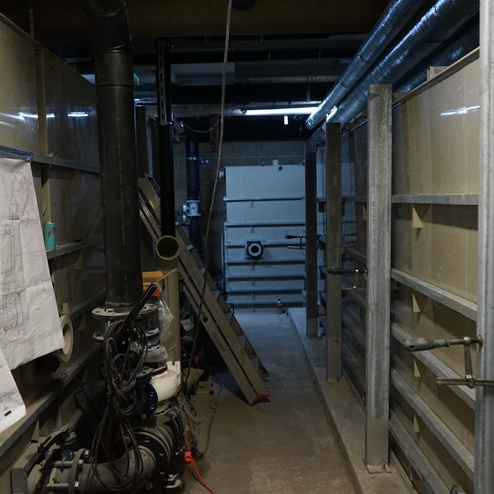 Langsamer, schmaler Gang zwischen W&auml;nden mit Rohren, einem Plan auf Papier und Werkzeugen, beleuchtet von Deckenlichtern.