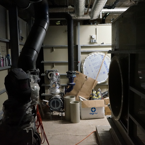 Dunkler Raum mit Rohren, Pumpen und Kartons, darunter ein gro&szlig;es Rohr und diverse Materialien auf dem Boden.