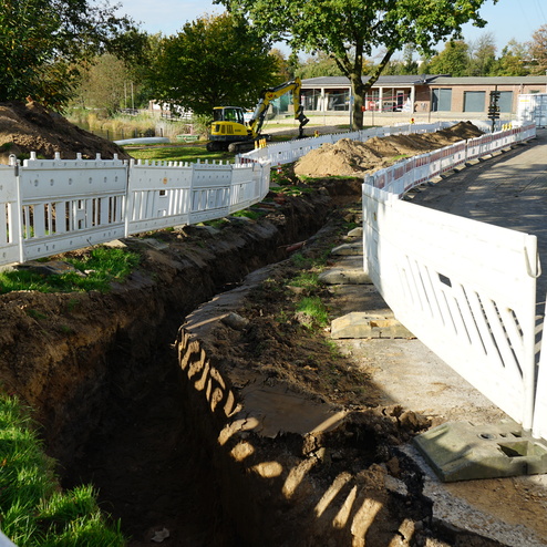 Baustelle mit einem tiefen Graben, umgeben von wei&szlig;en Baustellenabsperrungen und B&auml;umen im Hintergrund.
