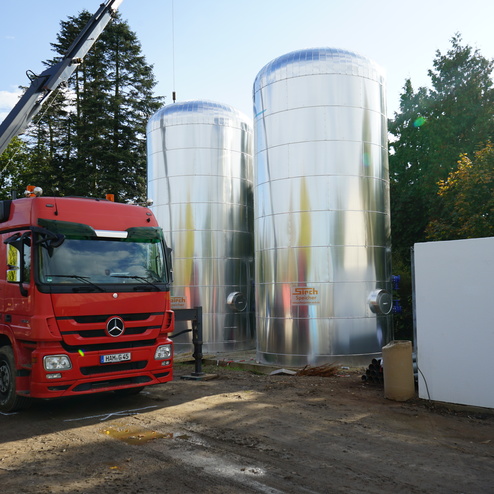 Roten Lkw mit Kran hebt einen silbernen Tank auf Baustelle zwischen zwei gro&szlig;en Wassertanks und B&auml;umen im Hintergrund.
