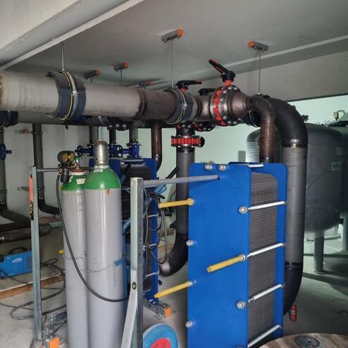 Anlage mit Rohren, Ventilen und zwei Gasflaschen in einem technischen Raum. Rechts ein blauer Wärmetauscher sichtbar.