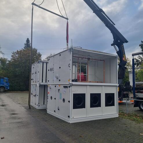 Kran hebt einen großen weißen Modulcontainer auf einem Pflasterplatz, umgeben von Bäumen und Fahrzeugen.