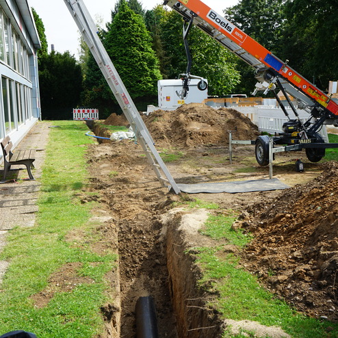 Baugel&auml;nde mit einer Grube, einer Baumaschine und Erdarbeiten entlang eines Gehwegs neben einem Geb&auml;ude.