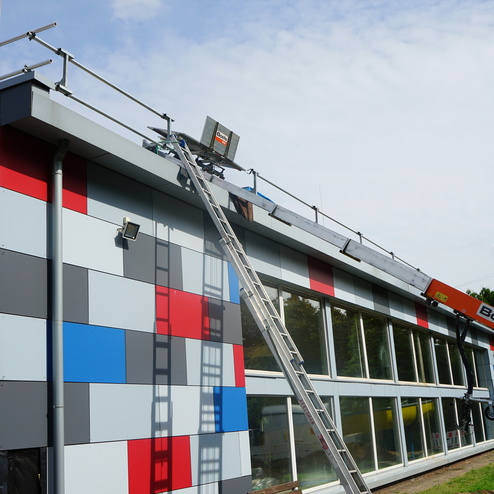 Blick auf ein Geb&auml;ude mit bunten Fassadenelementen, einer Leiter und einem Gelenkarmarbeitsb&uuml;hne zur Dacharbeit.